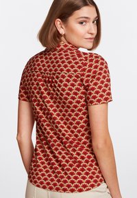 Camisa de manga corta con un patrón floral rojo y verde, hecha de tela suave con un dobladillo redondeado y un sutil detalle de pliegue en la parte posterior.