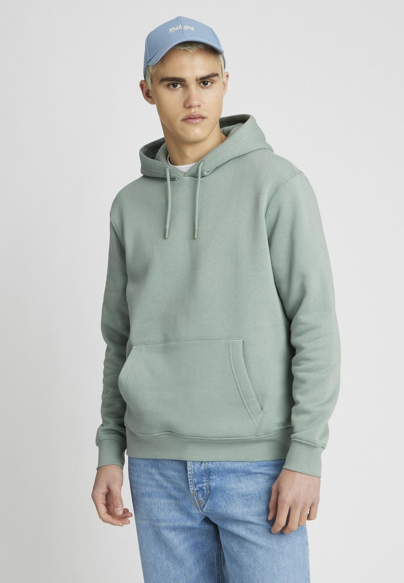 Haut à capuchon vert clair avec cordons, poche avant et poignets côtelés. Porté avec une casquette de baseball bleu clair et un jeans en denim.