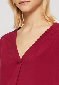 Blouse bordeaux avec un col en V, une texture douce et un léger drapé subtil à l'avant, mettant en valeur un style simple et élégant.
