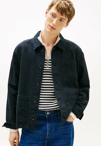 Marineblauwe jas met een knoopsluiting aan de voorkant, twee zakken en een kraagontwerp. Daaronder een gestreept shirt met contrasterende kleuren.