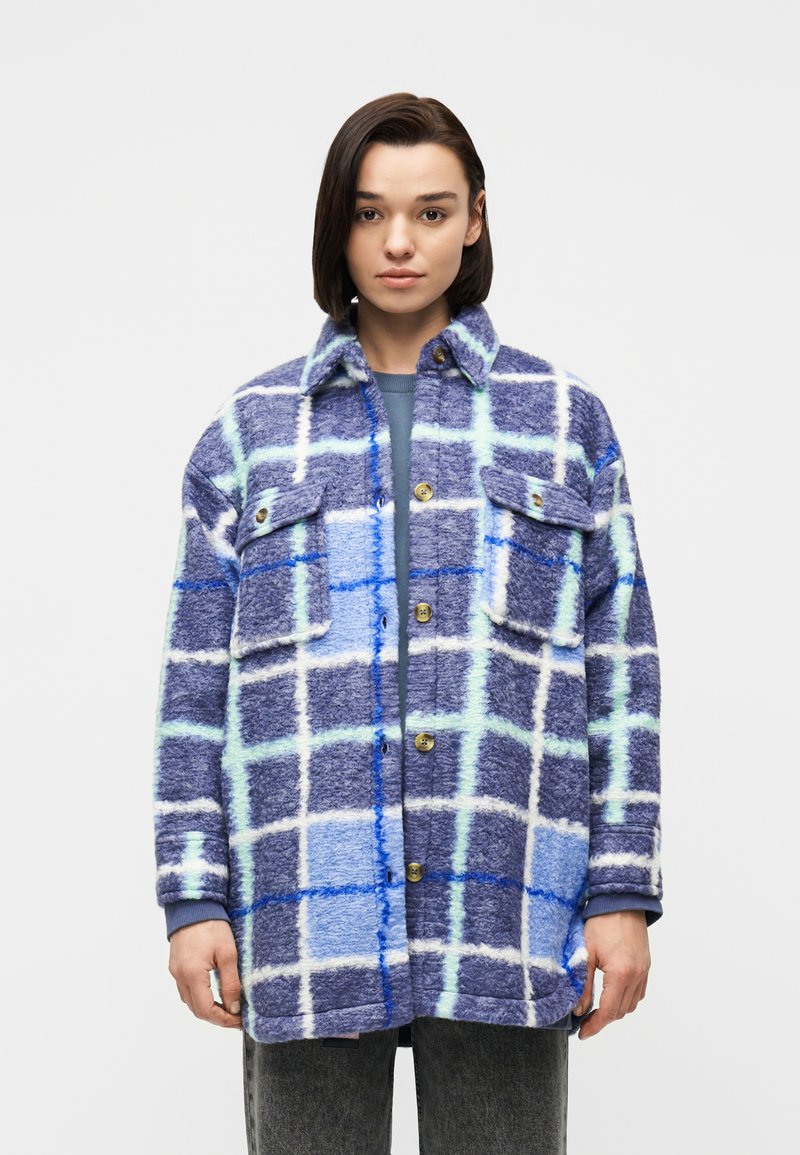 Νεαρό άτομο που φορά ένα μπλε και λευκό παλτό με κουμπιά και καρό σχέδιο, με μπροστινές τσέπες, στέκεται μπροστά σε έναν απλό ανοιχτό γκρι φόντο.