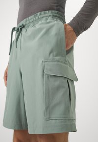 Šviesiai žali krovininiai šortai, pagaminti iš minkštos medžiagos, su raišteliu juosmens, šoninėmis kišenėmis ir uždara kišene kairiajame šlaunyje.