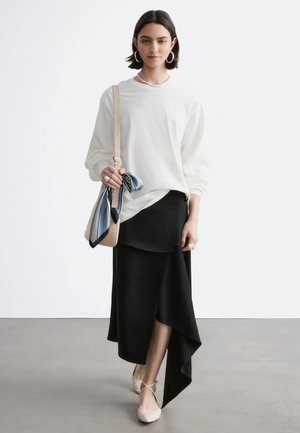 Mujer con blusa blanca de manga larga y falda negra asimétrica, llevando un bolso beige con pañuelo azul, caminando sobre suelo gris frente a una pared blanca.