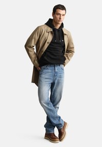 Tan trenchcoat over en sort hættetrøje, kombineret med lyseblå jeans og brune læderstøvler med en kraftig sål.