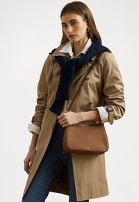 Brun läderaxelväska med en slät textur och guld-logotypdetalj, matchad med en beige trench coat och en marinblå stickad scarf.