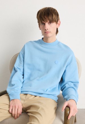Light blue sweatshirt with a crew neckline, ribbed cuffs, and a small embossed logo. Worn with beige pants, seated on a chair.