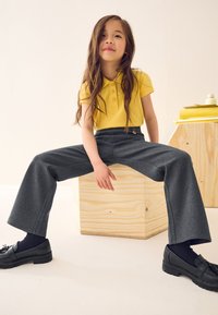 Kollane polo-särk, millel on kolm nööpi, hallid laiade säärtega püksid, mustad loafersid ja pikad pruunid juuksed. Istuvas asendis puitkastil.