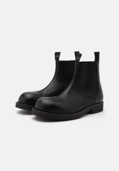 Une paire de bottines Chelsea en cuir noir à bout rond, à talons bas et à languettes, présentée sur un fond clair et uni.