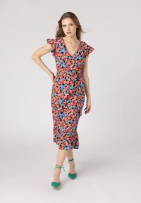 Robe portefeuille à fleurs avec un fond noir et des fleurs roses, bleues et vertes. Manches courtes et ceinture à nouer à la taille. Associée à des talons verts.