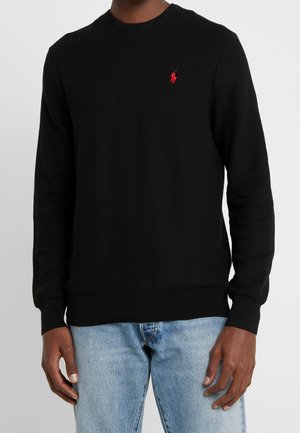 Mann trägt schwarzen, strukturierten Langarmpullover mit rotem, gesticktem Logo auf der Brust und hellblaue Jeans aus Denim, steht mit entspannten Händen.