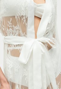 Διάφανη λευκή δαντελένια ρόμπα με floral σχέδια, δεμένη στη μέση πάνω από ένα λευκό σουτιέν, που δείχνει το στομάχι και μερική όψη του άνω μέρους του σώματος.