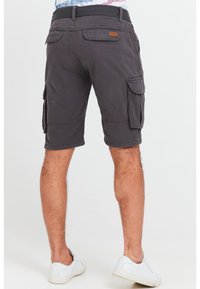 Man wearing dark gray cargo shorts with flap pockets and white sneakers, standing against a plain light background.