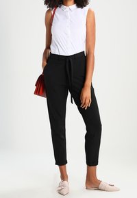 Pantalons noirs ajustés avec une taille nouée, associés à une chemise blanche à col. Complétés par des chaussures roses à enfiler et un sac à main rouge.