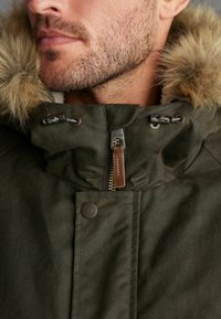Olivově zelená parka s kapucí podšitou kožešinou, zipem a patentkami. Obsahuje odolnou tkaninu a bronzové akcenty na kovových součástech.