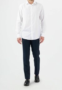 Bílá košile s dlouhým rukávem na knoflíky, kombinovaná s tmavě modrými kalhotami s tenkými pruhy a černými koženými botami, stojící před jednobarevným pozadím.