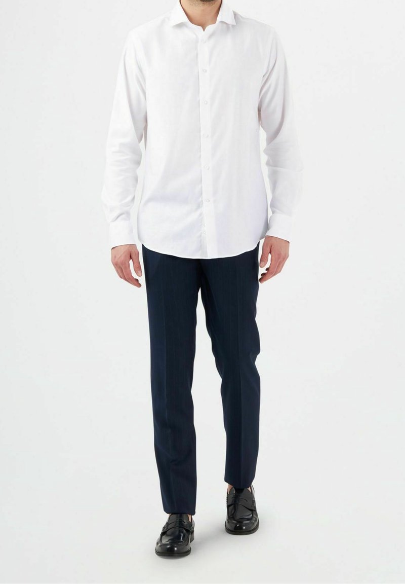 Bílá košile s dlouhým rukávem na knoflíky, kombinovaná s tmavě modrými kalhotami s tenkými pruhy a černými koženými botami, stojící před jednobarevným pozadím.