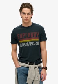 Sort bomulds t-shirt med fed rød "SUPERDRY" tekst og vandrette striber i gul og grå, kombineret med blå denim jeans og en grå sweater bundet om taljen.