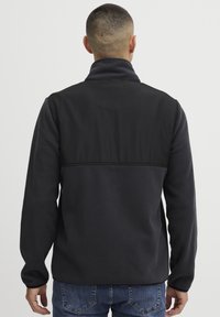 Chaqueta negra con cuerpo de forro polar y un tejido superior suave. Presenta un collar alto y mangas largas, combinada con jeans azules.