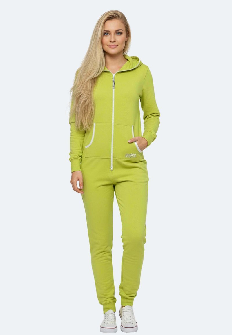 Jumpsuit verde con zip, dotato di tasche frontali, dettagli bianchi e design aderente. Il modello indossa sneakers bianche. Sullo sfondo un colore neutro.