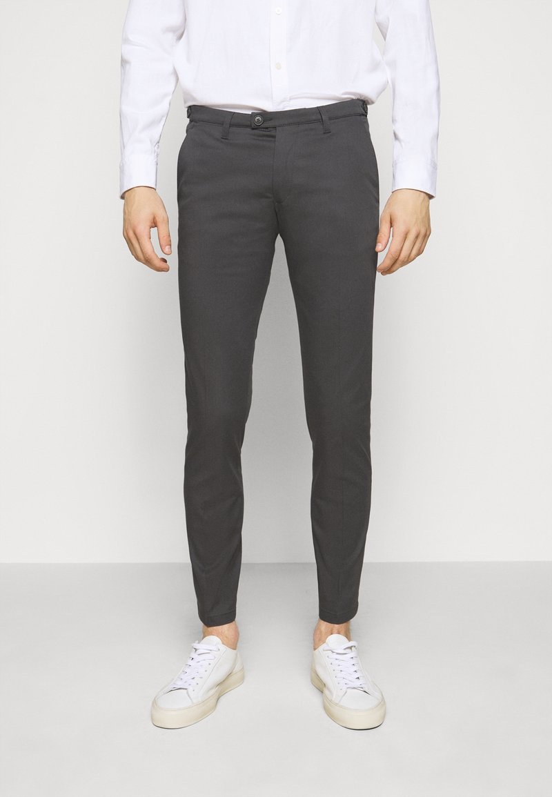 Homme portant un pantalon gris foncé ajusté, une chemise blanche boutonnée et des baskets blanches, debout devant un fond clair uni.