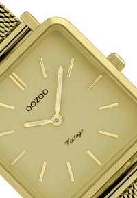 Montre rectangulaire dorée avec un bracelet en maille, dotée d'un cadran minimaliste, d'index des heures dorés et d'une petite couronne bleue sur le côté.