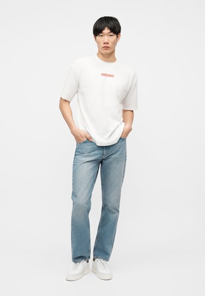 Jeune homme debout, les mains dans les poches, portant un T-shirt blanc avec le texte "WRONG", un jean bleu clair et des baskets blanches sur un fond uni.