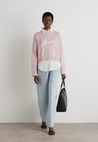 Lys rosa strikkegenser med "Boss"-tekst, hvit skjorte med krage under, lyseblå vide jeans og svart håndveske, stående på nøytral gulv.