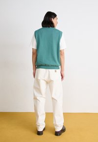 Gilet verde lavorato a maglia sopra una maglietta bianca, abbinato a pantaloni larghi color crema e scarpe nere con accenti viola.