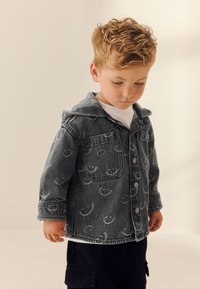 Giacca di jeans con cappuccio, grigia con motivi di faccine sorridenti, chiusura con bottoni e due tasche sul petto. Sotto si vede una camicia bianca.