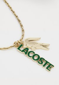 Collier en chaîne dorée avec un logo crocodile Lacoste vert et or, ainsi que le texte "LACOSTE", doté d'une finition lisse et d'une lettrage en relief.