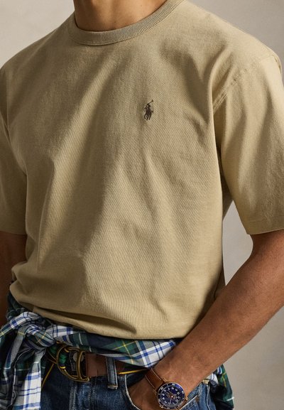 Homme portant une chemise beige à manches courtes avec un logo brodé, chemise à carreaux nouée à la taille, jean bleu et montre-bracelet au poignet gauche.