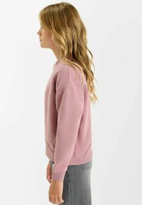 Roze sweatshirt van zacht materiaal, met een ronde halslijn en geribbelde manchetten, gedragen met grijze jeans, zijaanzicht.