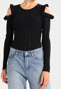 Zwarte gebreide top met een cold shoulder ontwerp, geribbelde textuur en ruches; gecombineerd met lichtblauwe distressed denim jeans.