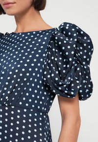 Robe bleu marine avec manches bouffantes, ornée d'un motif à pois bleu clair. Tissu lisse avec un corsage ajusté et une jupe fluide.