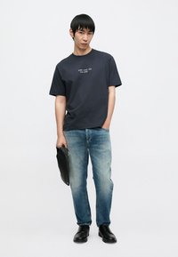 T-shirt en coton bleu foncé avec texte blanc, jean bleu clair usé, et chaussures noires. Une personne tient une pochette noire. Fond neutre.