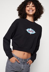 Felpa nera corta con maniche lunghe, caratterizzata da un logo "silverTab" multicolore. Abbinata a jeans a vita alta di un grigio chiaro.