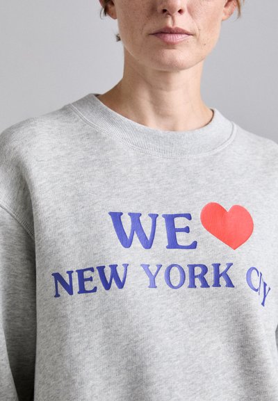 Sivá mikina s okrúhlym výstrihom, na ktorej sa nachádza výrazný text "MILUJEME NEW YORK CITY" v purpurovej a červenej farbe. Mäkko bavlnený materiál, voľný strih.