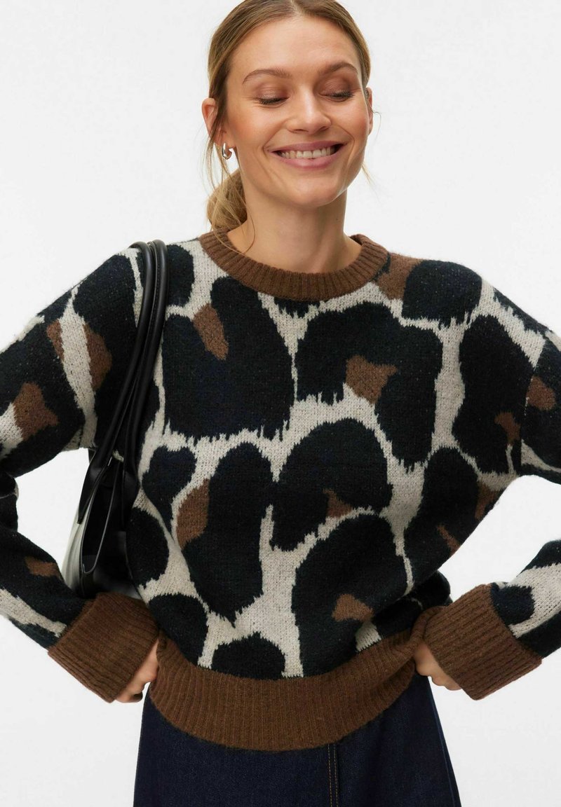 Femme souriante portant un pull marron et noir à imprimé animal avec poignets et ourlet côtelés, mains sur les hanches, portant un sac noir à bandoulière.