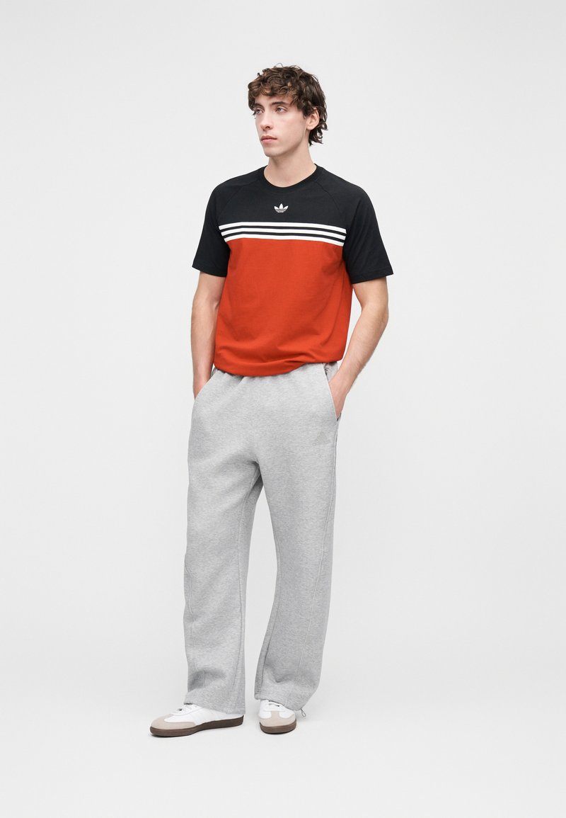 T-shirt à manches courtes rouge et noir avec des rayures blanches, assorti à un pantalon de survêtement gris et des chaussures de sport blanches. Design simple et sportif.