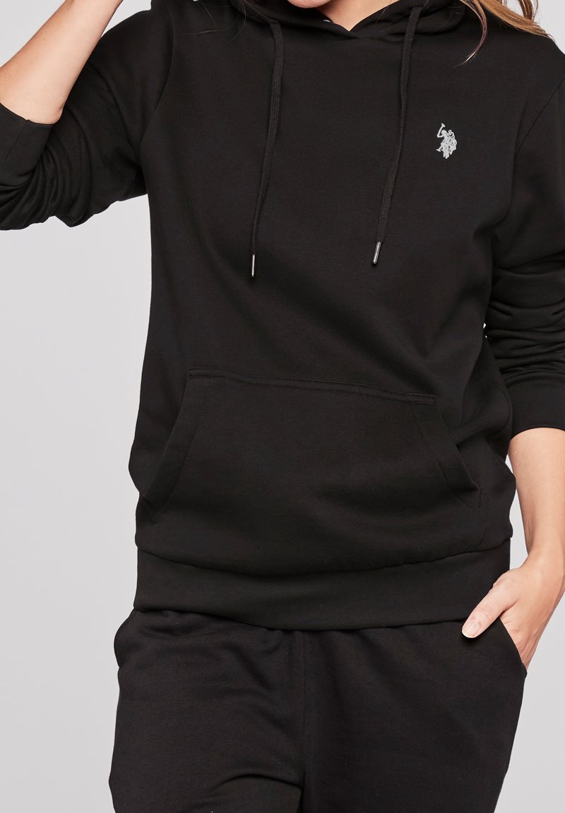 Personne portant un sweat à capuche noir avec poche avant et cordons, avec un petit logo blanc sur le côté gauche de la poitrine, main dans la poche.