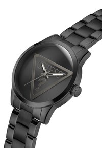 Orologio analogico nero con quadrante triangolare e superficie testurizzata, presenta il logo "GUESS" e un design a punto interrogativo. Bracciale a maglie metalliche.