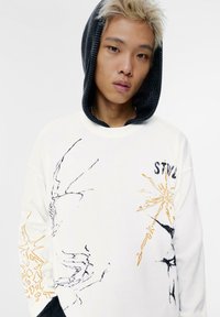 Sweatshirt alb cu designuri abstracte negre și portocalii, cu o glugă texturată neagră și o croială lejeră. "STYL" imprimat pe față.