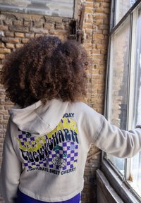 Korte hoodie van beige stof met een gekleurde grafische print met dobbelstenen en bloemen op de achterkant. Tekst luidt "Lucky Charm."