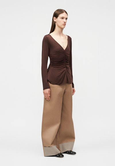 Γυναίκα που φοράει καφέ μακρυμάνικο τοπ με ρυτίδες, φαρδιά μπεζ παντελόνια με καρό μανσέτες και μαύρα μυτερά παπούτσια, στέκεται μπροστά σε απλό φόντο.