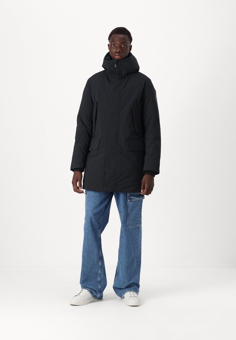 Parka nera con colletto alto, cerniera frontale, due ampi flap per le tasche e cappuccio removibile, indossata sopra jeans a gamba larga di un blu chiaro.
