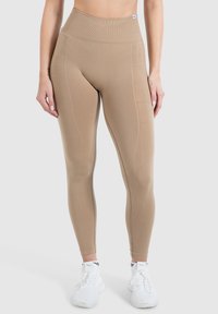 Beige højtaljede leggings lavet af strækbart stof, med teksturerede paneler og syninger, parret med hvide træningssko.