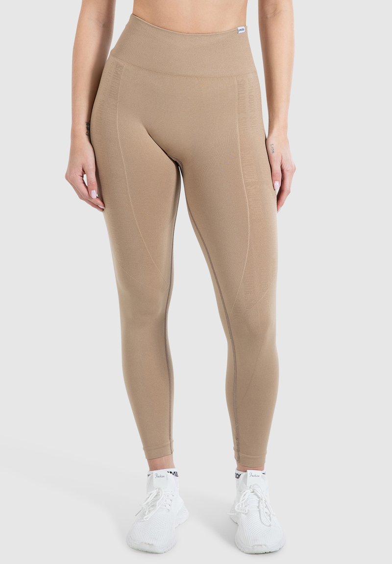 Beige højtaljede leggings lavet af strækbart stof, med teksturerede paneler og syninger, parret med hvide træningssko.