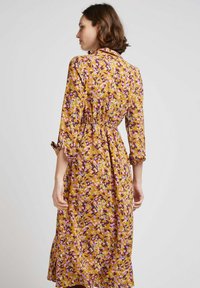 Robe midi florale en jaune moutarde avec des accents violets et blancs, présentant une taille cintrée, des manches longues et un ourlet volé.