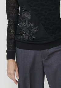 La mano di una donna poggia su un maglione scuro e lavorato a maglia, decorato con motivi floreali, abbinato a dei pantaloni grigi su misura.