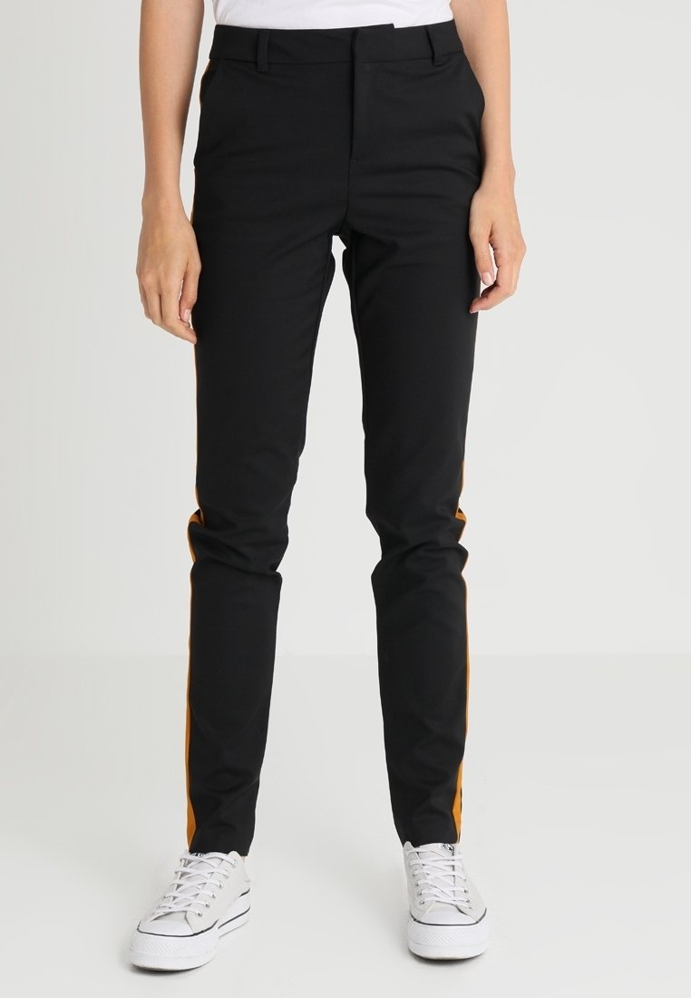 Personne portant un pantalon noir slim avec des bandes latérales orange et des baskets blanches, debout devant un fond clair uni.